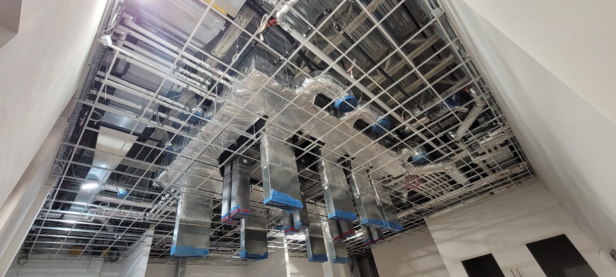Open T-bar ceiling grid with exposed overhead ductwork and services during a Vancouver commercial interior installation. Project archive photo supplied by Mason McKenzie / Rambo Wall & Ceiling Limited; installation date not confirmed. Open T-bar ceiling grid with exposed overhead ductwork and services during a Vancouver commercial interior installation. Project archive photo supplied by Mason McKenzie / Rambo Wall & Ceiling Limited; installation date not confirmed.